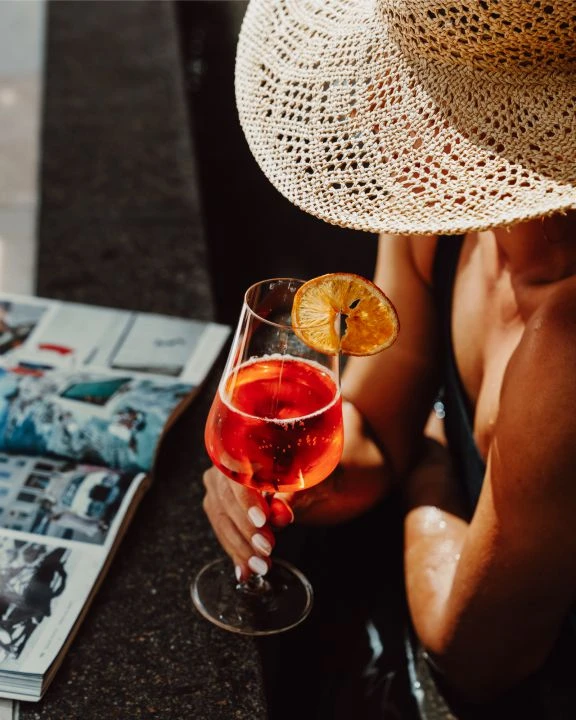 Frau mit Strohhut am Poolrand mit Getränk und Zeitung