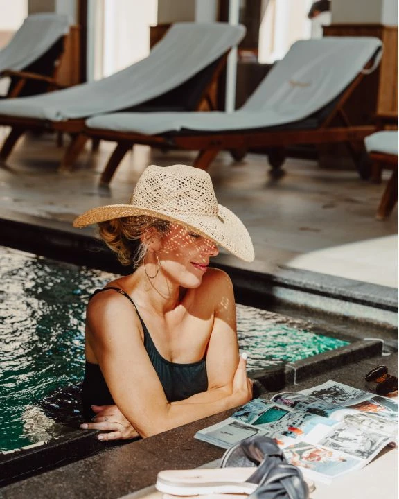 Frau mit Strohhut am Poolrand mit Zeitung