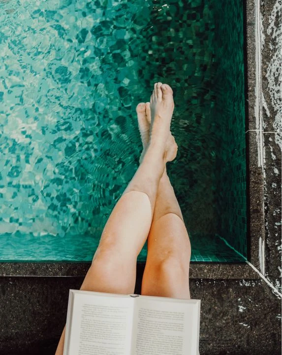 Unsere Philosophie 2 Frau sitzt am Poolrand mit Buch und Beine im Wasser