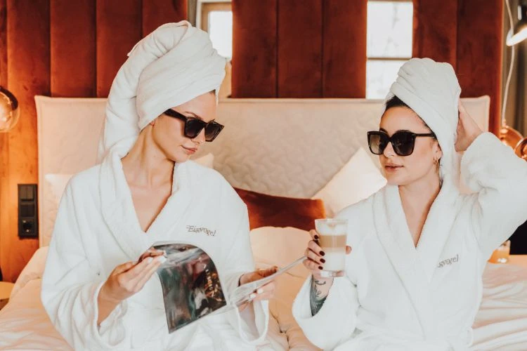 Zwei Freundinnen sitzen mit Bademantel, Turban und Sonnenbrille am Bettrand