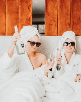Zwei Freundinnen trinken Sekt im Bett mit Bademantel, Turban und Sonnebrille